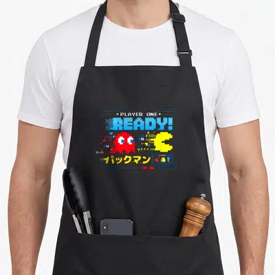 Tablier de cuisine noir orné d'un graphisme pixelisé jaune et rouge avec le texte Player One Ready en bleu.