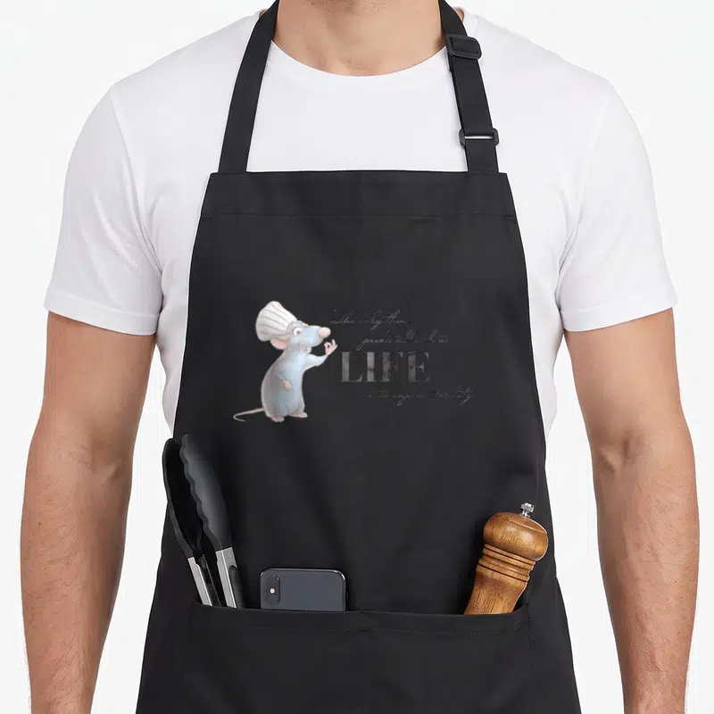 Tablier de cuisine noir avec Remy en toque blanche et texte calligraphié sur fond gris uni.