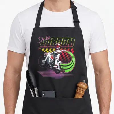 Tablier de cuisine noir avec illustration colorée d'un cascadeur sur moto et le texte Duke Caboom en lettres néon.
