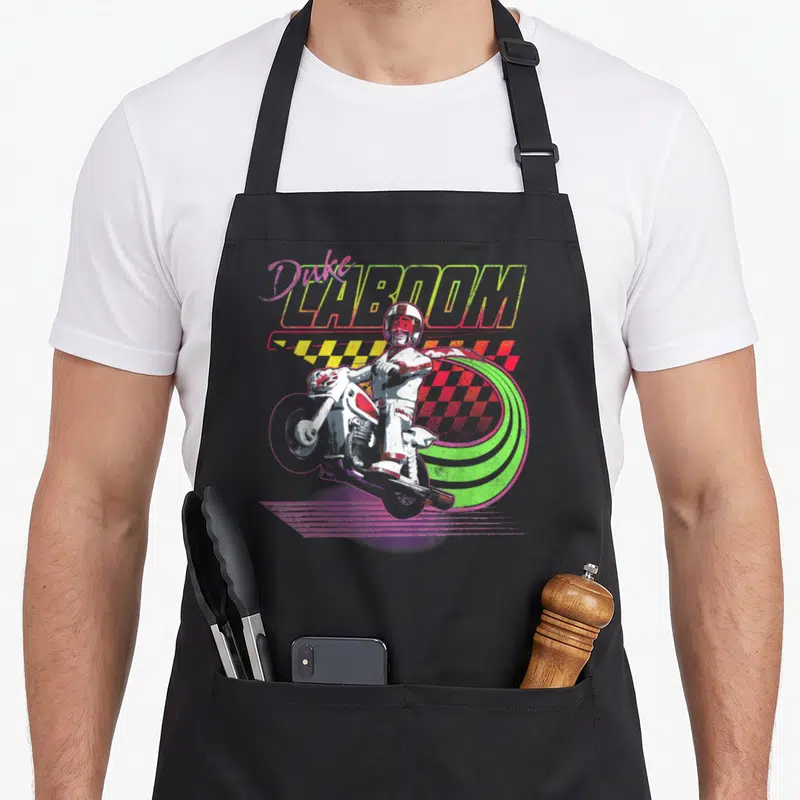 Tablier de cuisine noir avec illustration colorée d'un cascadeur sur moto et le texte Duke Caboom en lettres néon.
