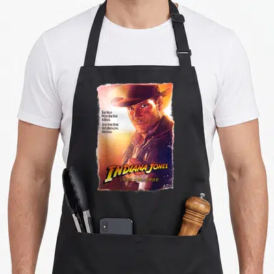 Tablier de cuisine noir avec une illustration colorée représentant un aventurier au chapeau et le titre du film en lettres jaunes.