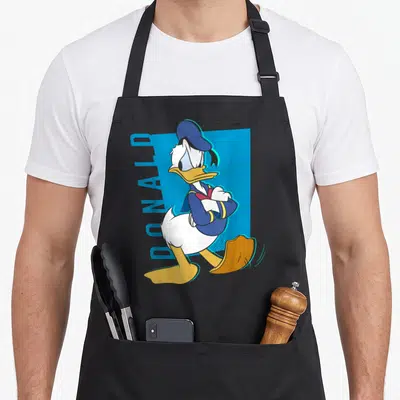 Tablier de cuisine noir avec illustration colorée de Donald Duck aux bras croisés et texte vertical DONALD sur fond bleu.