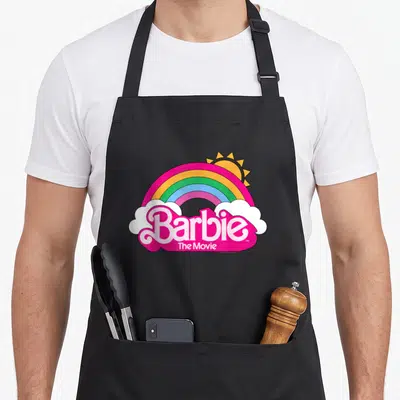 Tablier de cuisine noir avec un arc-en-ciel rose et les mots Barbie The Movie en lettres roses vives sur des nuages blancs.
