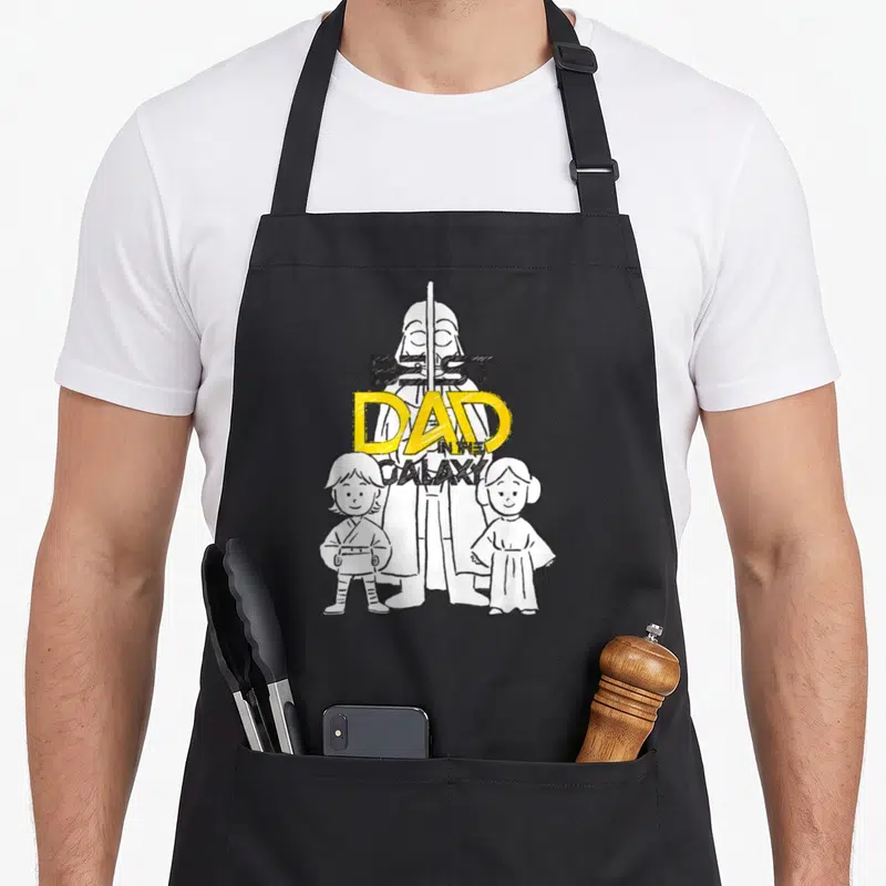 Tablier de cuisine noir avec illustration Darth Vader, Luke et Leia et texte Best Dad in the Galaxy en jaune et noir.