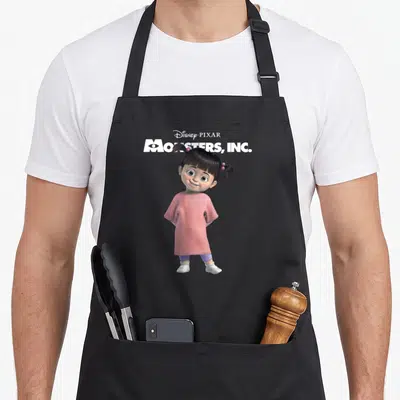 Tablier de cuisine noir avec illustration de Boo en robe rose et titre Monsters Inc en haut.