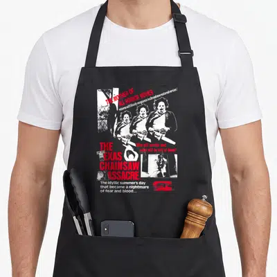 Tablier de cuisine noir avec illustration blanche et rouge représentant des personnages armés de tronçonneuses et du titre du film en typographie vintage.