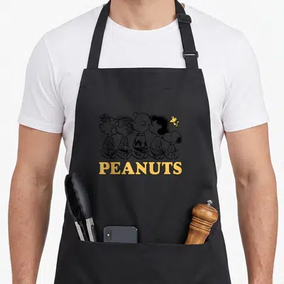 Tablier de cuisine noir avec illustration Snoopy Lucy Linus Charlie Sally Woodstock Walking et logo Peanuts jaune.