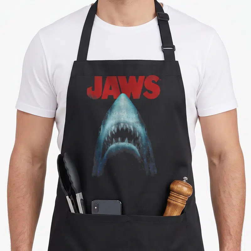 Tablier de cuisine noir avec illustration d'un grand requin blanc aux dents visibles et le mot JAWS en lettres rouges au-dessus.