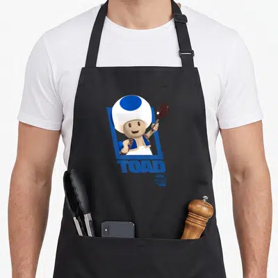 Tablier de cuisine noir avec Toad tenant une cuillère sous un cadre bleu et le mot TOAD imprimé en dessous.