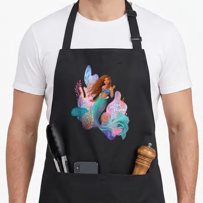 Tablier de cuisine noir avec une sirène souriante aux cheveux longs, entourée de coraux multicolores et du texte find your voice en blanc.