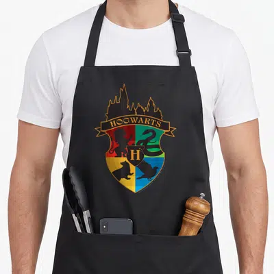 Tablier de cuisine noir avec le blason coloré de Poudlard et les silhouettes des maisons.