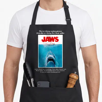 Tablier de cuisine noir avec reproduction du poster de Jaws montrant un grand requin et une nageuse.