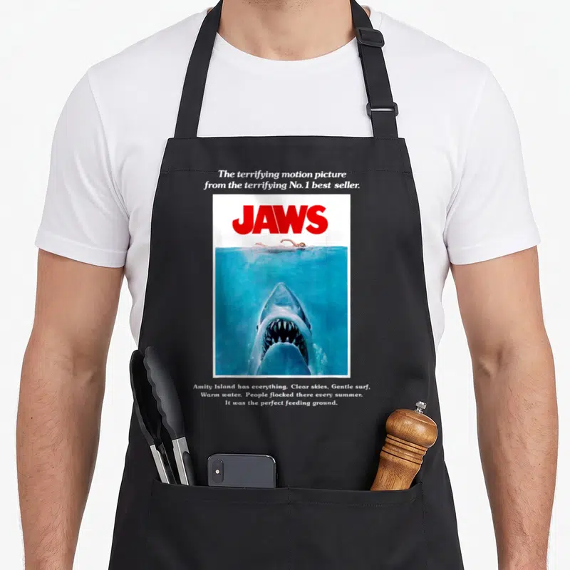 Tablier de cuisine noir avec reproduction du poster de Jaws montrant un grand requin et une nageuse.