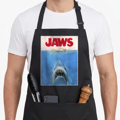 Tablier de cuisine noir avec la pochette du film Jaws montrant un requin et une nageuse.