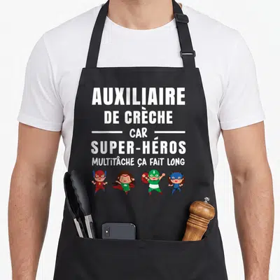 Tablier de cuisine noir avec texte blanc auxiliaire de crèche et quatre petits super-héros en dessous.