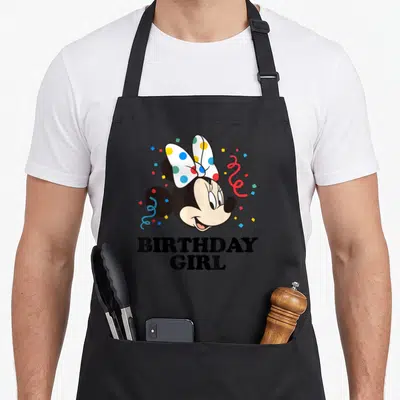 Tablier de cuisine noir avec lillustration de Minnie Mouse anniversaire Girl entourée de confettis et de guirlandes festives.