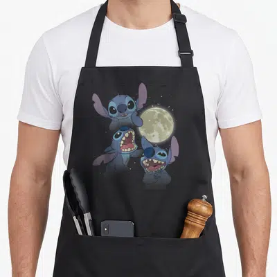 Tablier de cuisine noir avec trois illustrations de Stitch sous une lune jaune pâle et des étoiles blanches dispersées sur fond sombre.