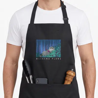 Tablier de cuisine noir avec Meeko allongé dans la forêt et le texte WEEKEND PLANS imprimé en blanc.