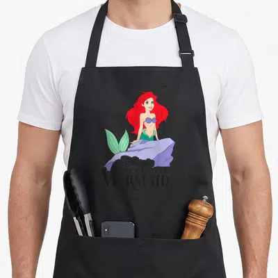 Tablier de cuisine noir avec Ariel la sirène aux cheveux roux et le titre du film classique en typographie noire distinctive.