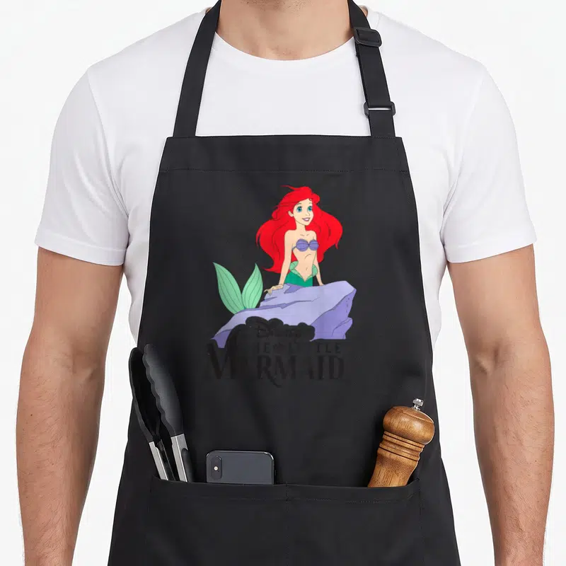 Tablier de cuisine noir avec Ariel la sirène aux cheveux roux et le titre du film classique en typographie noire distinctive.