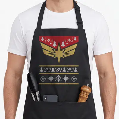 Tablier de cuisine noir avec motif pull de Noël Captain Marvel aigle doré et sapins blancs sur fond rouge.
