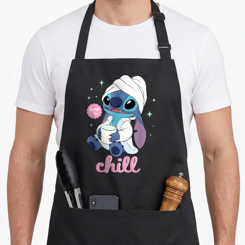 Tablier de cuisine noir avec Stitch enveloppé dans une serviette tenant une tasse blanche.