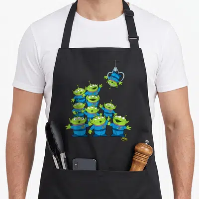 Tablier de cuisine noir avec un groupe d'aliens verts et une griffe mécanique suspendue au-dessus.
