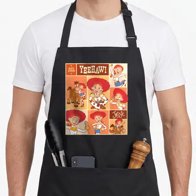 Tablier de cuisine noir avec un motif coloré représentant Jessie, Bullseye et le texte YEEHAW. en lettres capitales blanches.