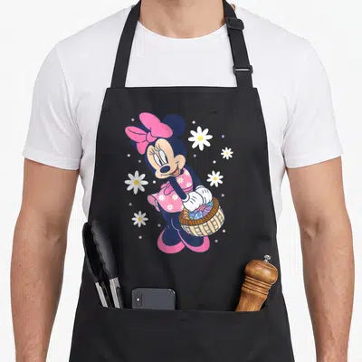 Tablier de cuisine noir avec Minnie Mouse tenant un panier d'œufs de Pâques entouré de fleurs blanches.