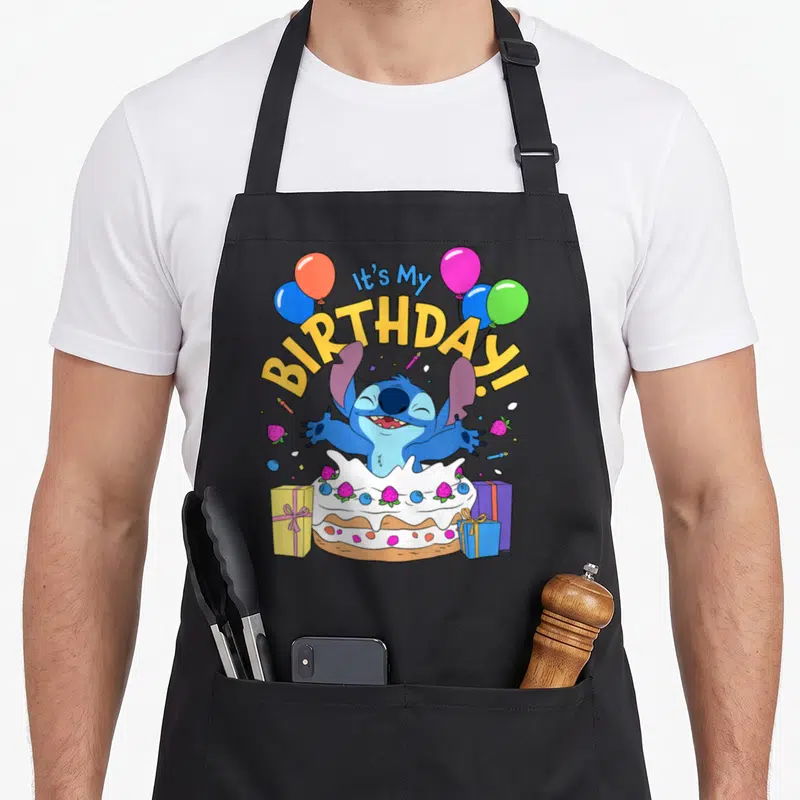 Tablier de cuisine noir avec Stitch sortant d'un gâteau d'anniversaire entouré de ballons et de cadeaux.