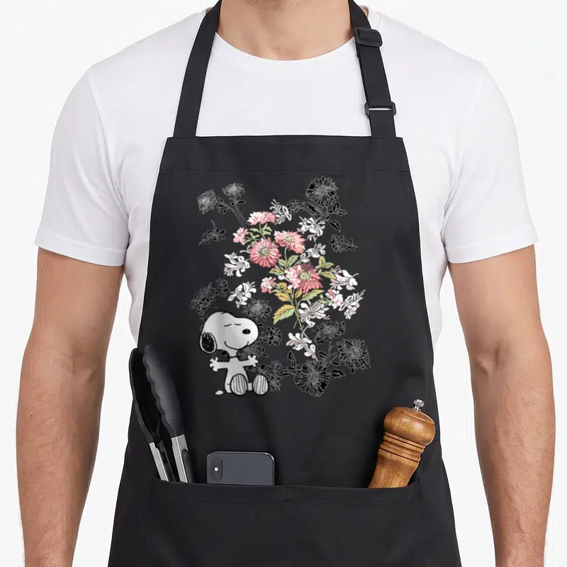 Tablier de cuisine noir avec Snoopy assis entouré de fleurs roses blanches et noires stylisées.