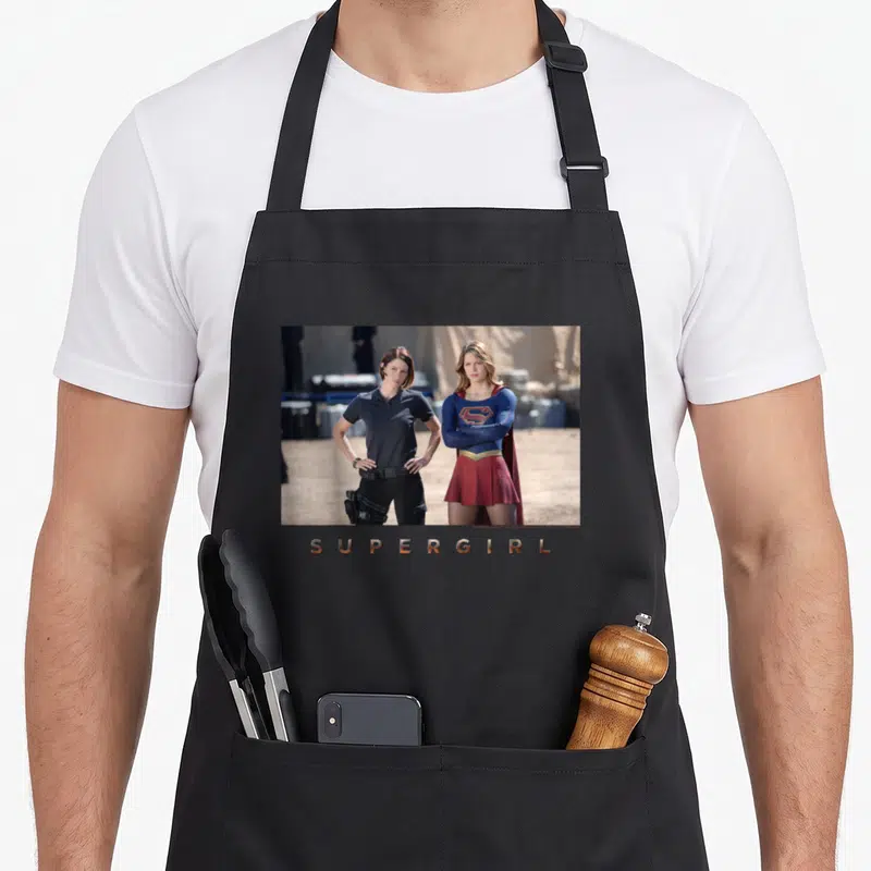 Tablier de cuisine noir avec photographie des personnages de Supergirl et le titre doré en bas.