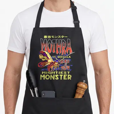 Tablier de cuisine noir avec illustration de Mothra et texte Mothra vs Godzilla Mightiest Monster en rouge et jaune.