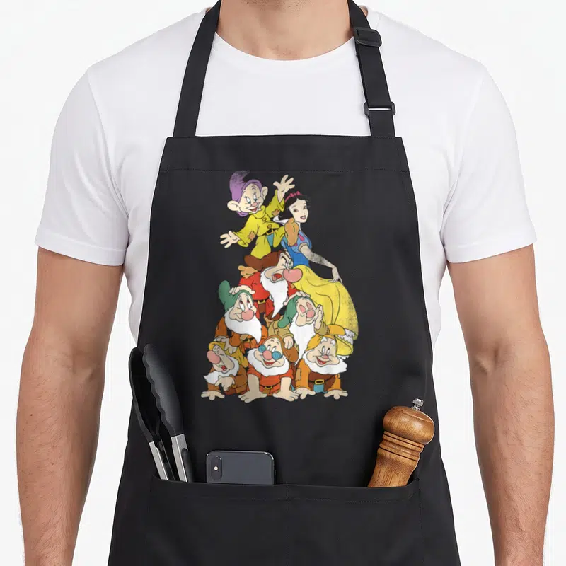 Tablier de cuisine noir avec une illustration colorée représentant Blanche-Neige et les sept nains formant une pyramide humaine sur le devant.