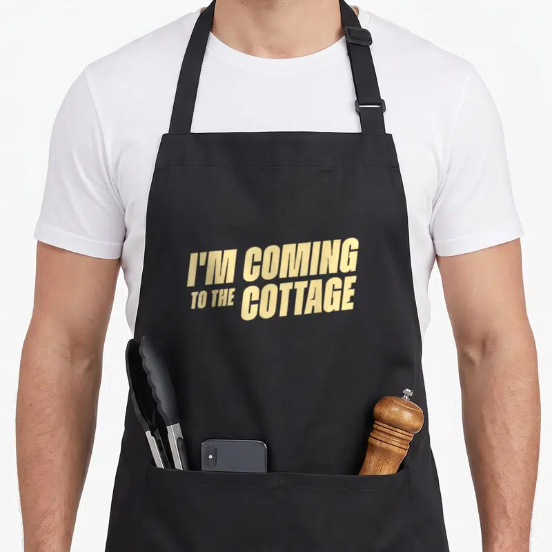 Tablier de cuisine noir avec l'inscription I'M COMING TO THE COTTAGE en typographie jaune diagonale.