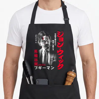 Tablier de cuisine noir avec portrait stylisé de John Wick et caractères japonais rouges sur fond gris.