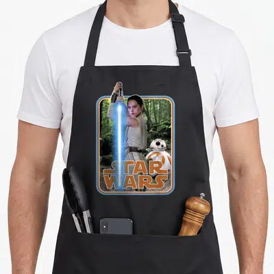 Tablier de cuisine noir avec illustration de Rey tenant une lightsaber bleue et le robot BB-8 devant un logo Star Wars orange.