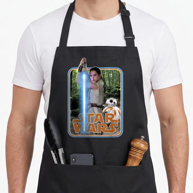 Tablier de cuisine noir avec illustration de Rey tenant une lightsaber bleue et le robot BB-8 devant un logo Star Wars orange.