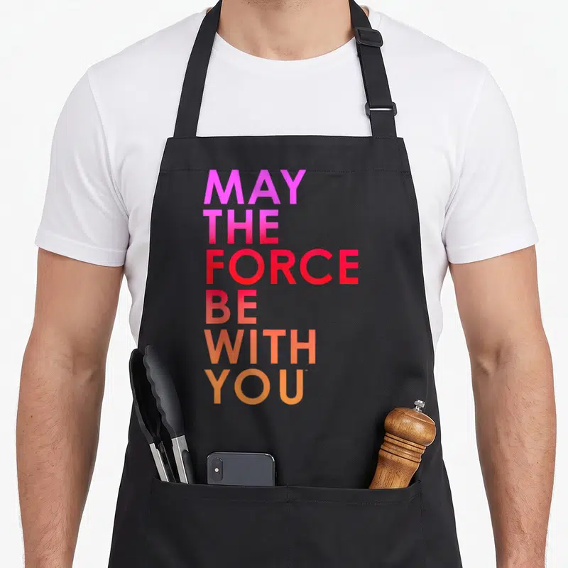 Tablier de cuisine noir avec l'inscription May the Force Be With You en lettres colorées verticales.