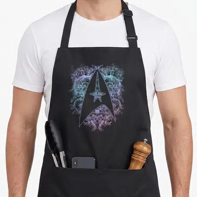 Tablier de cuisine noir avec un insigne Starfleet pastel orné de motifs végétaux complexes et détaillés.