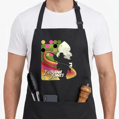 Tablier de cuisine noir avec silhouette Indiana Jones, fouet arc-en-ciel et texte vintage sur fond de pois colorés.