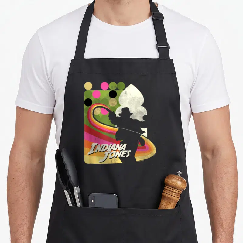 Tablier de cuisine noir avec silhouette Indiana Jones, fouet arc-en-ciel et texte vintage sur fond de pois colorés.
