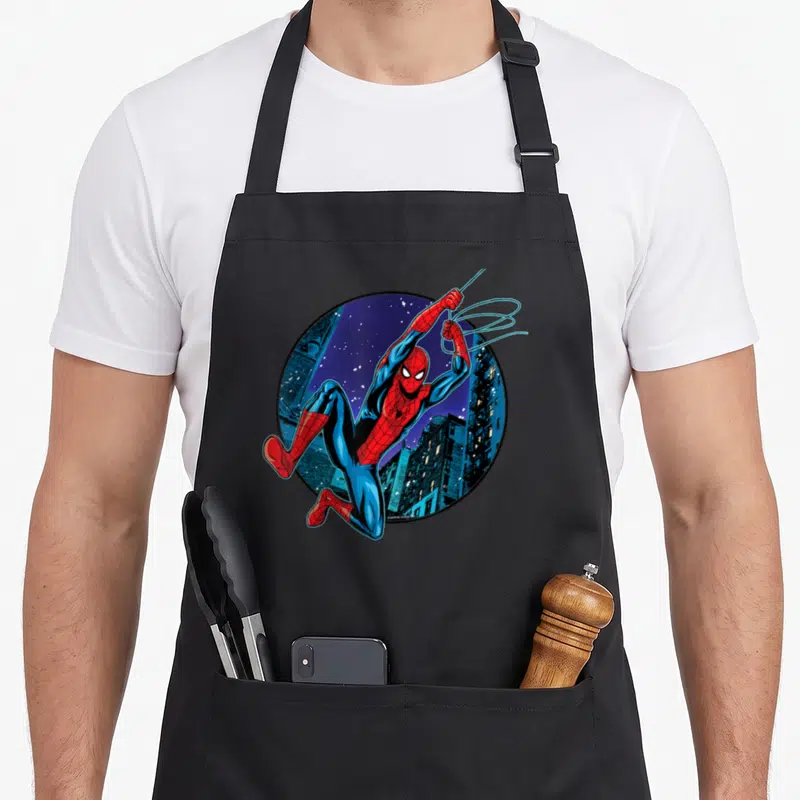 Tablier de cuisine noir avec Spider-Man en train de swinguer devant une ville nocturne stylisée.