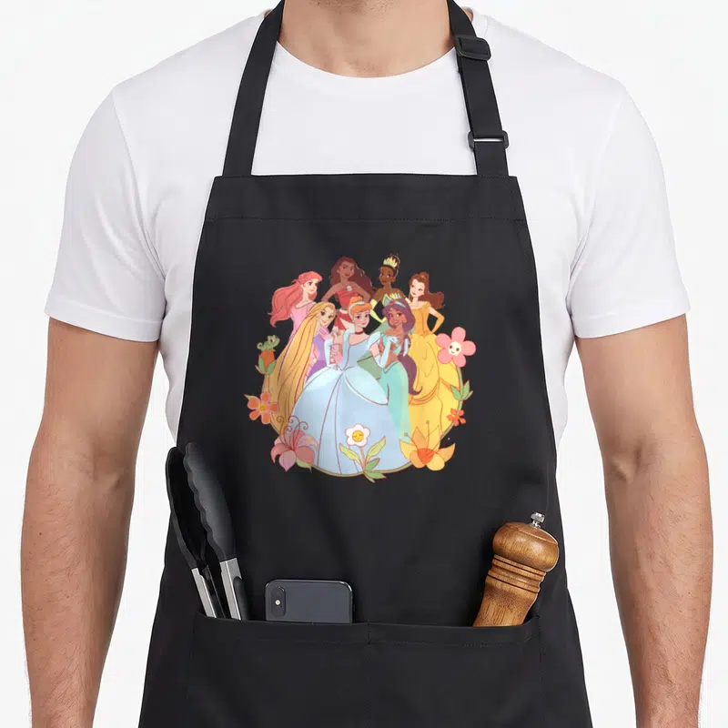 Tablier de cuisine noir avec des princesses en robes pastel entourées de fleurs colorées et d'un petit lézard.