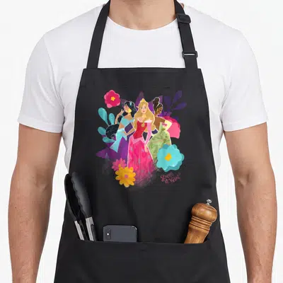 Tablier de cuisine noir avec illustration des princesses Jasmine, Aurora et Tiana parmi des fleurs roses et bleues.