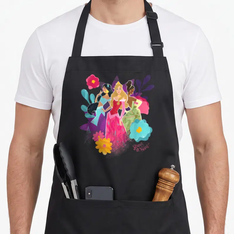 Tablier de cuisine noir avec illustration des princesses Jasmine, Aurora et Tiana parmi des fleurs roses et bleues.
