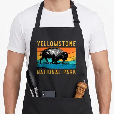 Tablier de cuisine noir avec illustration rétro d'un bison devant un ciel orange et bleu, texte Yellowstone National Park en jaune.