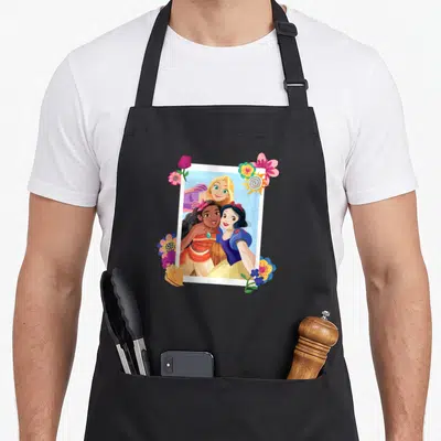 Tablier de cuisine noir avec Raiponce Moana et Snow White posant ensemble dans un cadre polaroid entouré de fleurs colorées.
