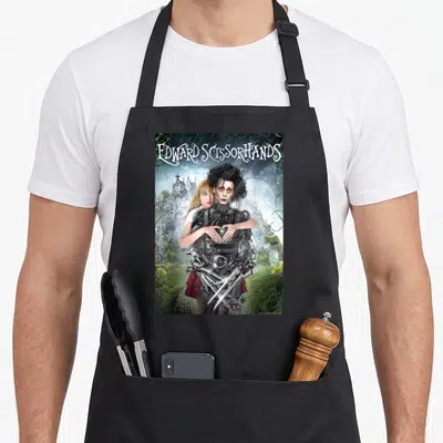 Tablier de cuisine noir avec une illustration d'Edward Scissorhands et Kim dans un jardin féerique sous la pleine lune.