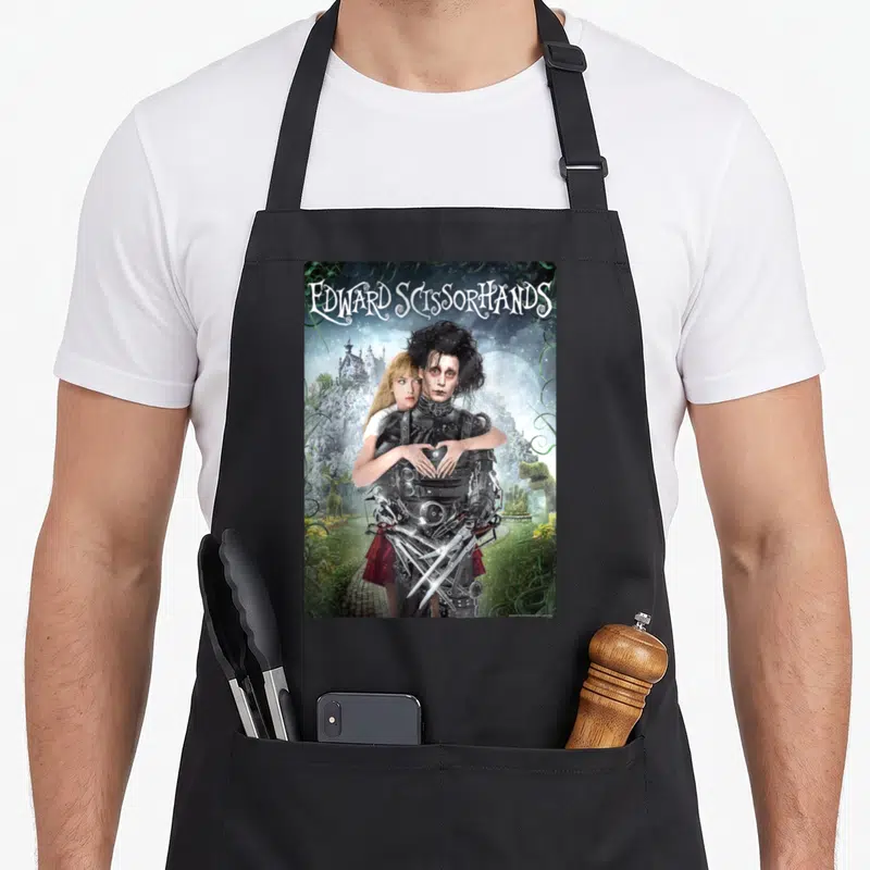 Tablier de cuisine noir avec une illustration d'Edward Scissorhands et Kim dans un jardin féerique sous la pleine lune.